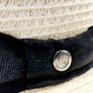 Close-up of Sbarià natural straw bombetta hat with spiral pin, metallic button, black band, and white woven straw detail.